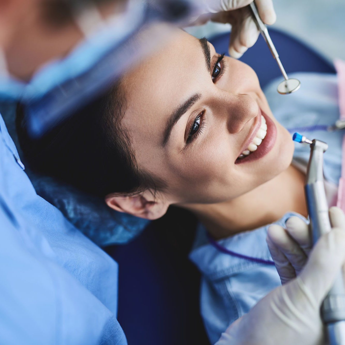 Close up portrait of charming woman sitting in dental chair while stomatologist holding polisher and mirror. Girl is smiling